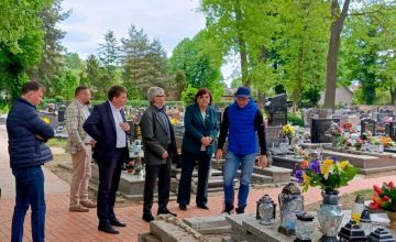 Sześć osób stojących na jednej z cmentarnych uliczek. Chodnik wykonany jest z czerwonej kostki. W oddali widać groby oraz zielone drzewa.